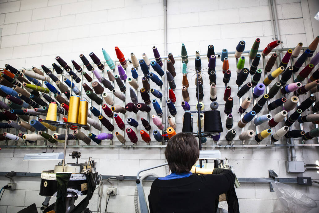 Yarns of different coloured thread mounted on a wall above a woman at a sewing machine