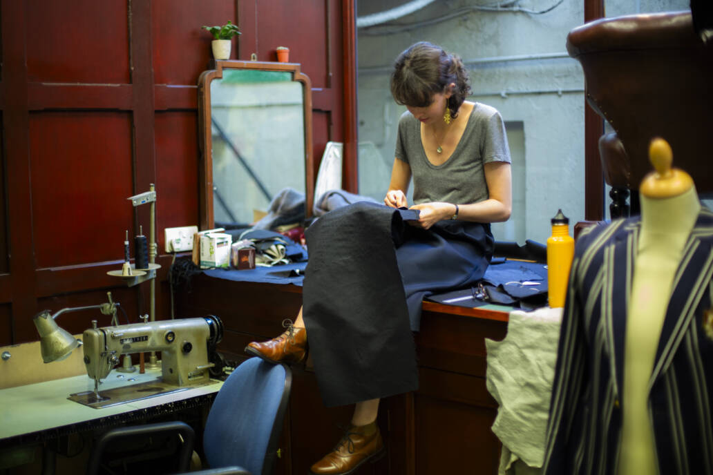 Woman sitting on a bench next to a window while stitching an item of clothing