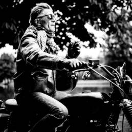 Black and white image of silver fox man in sunglasses riding a motorcycle one handed with lake and trees in the background having a cigarette