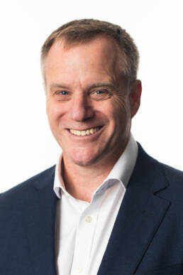 Colour head shot of a smiling man in a suit without a tie against a white background