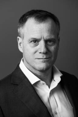 Black and white headshot of a man in a suit without a tie against a dark background